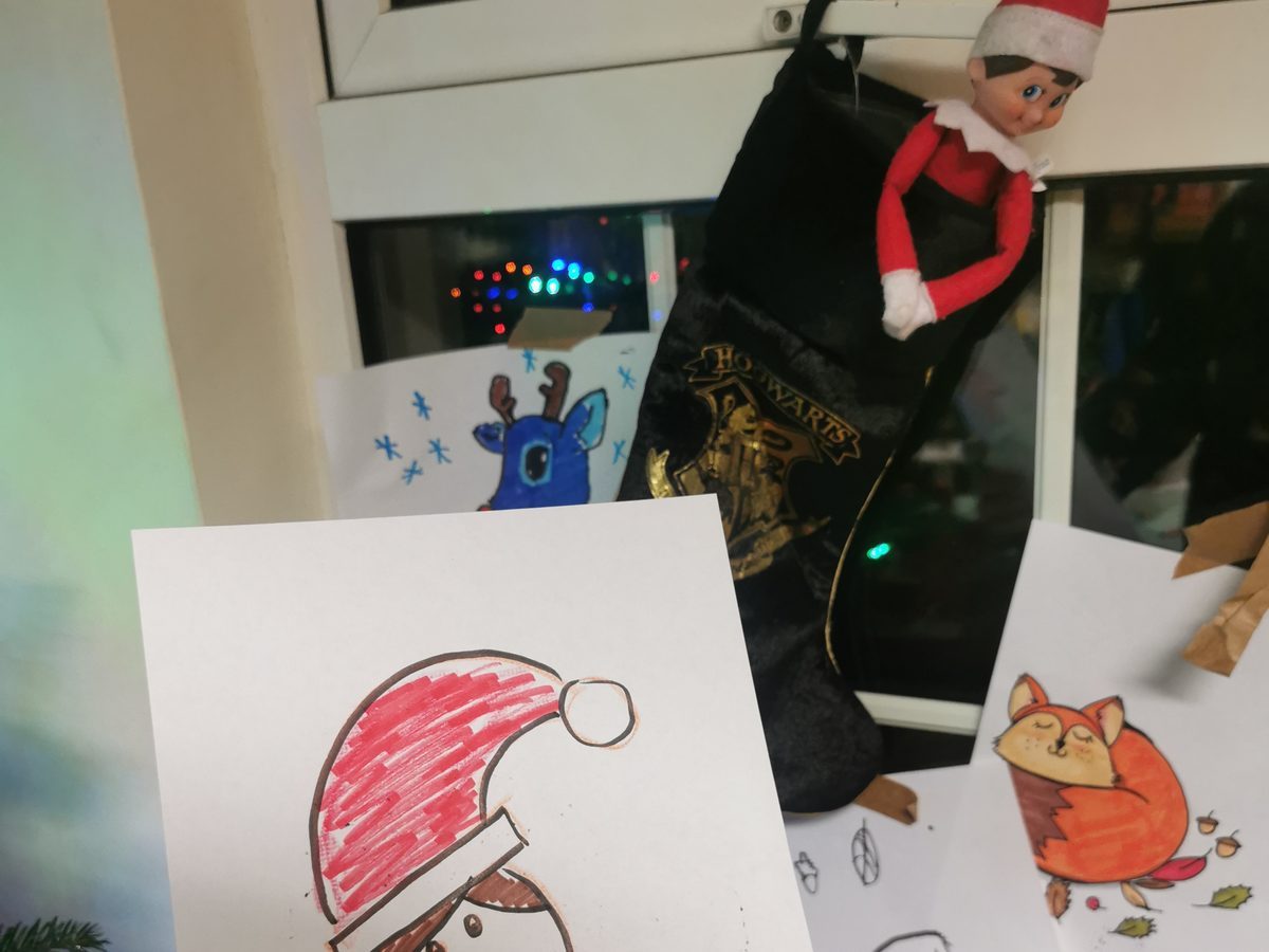 Elf at a small table with paper, markers, and a self-portrait drawing.