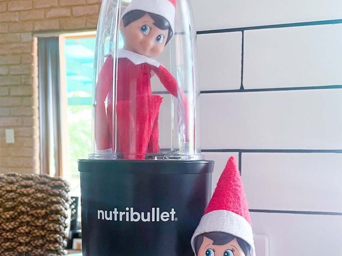 Two Christmas elves with one sitting inside blender creating mischievous scene.