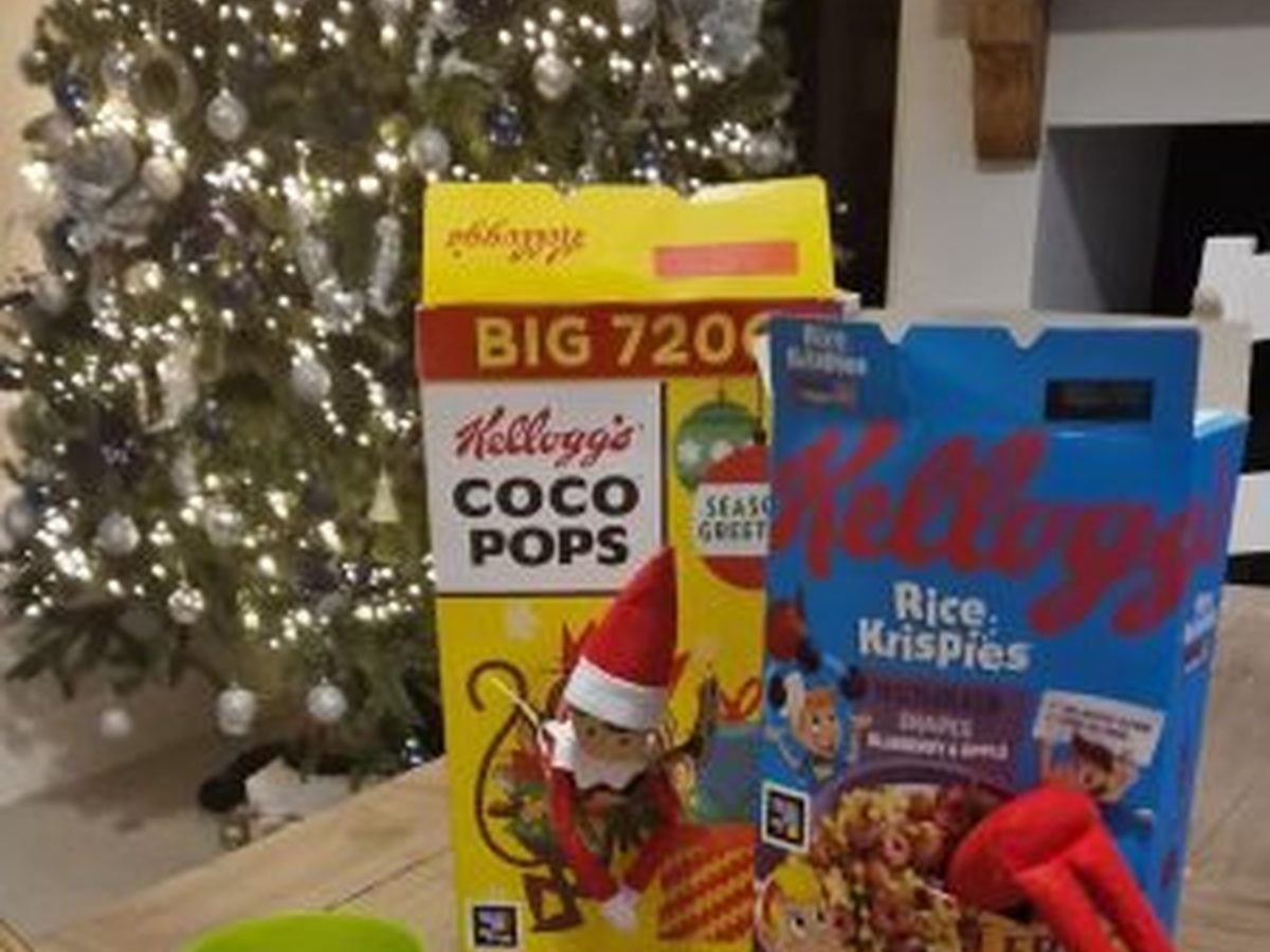 Elf perched in open cereal box with scattered cereal pieces on kitchen surface.