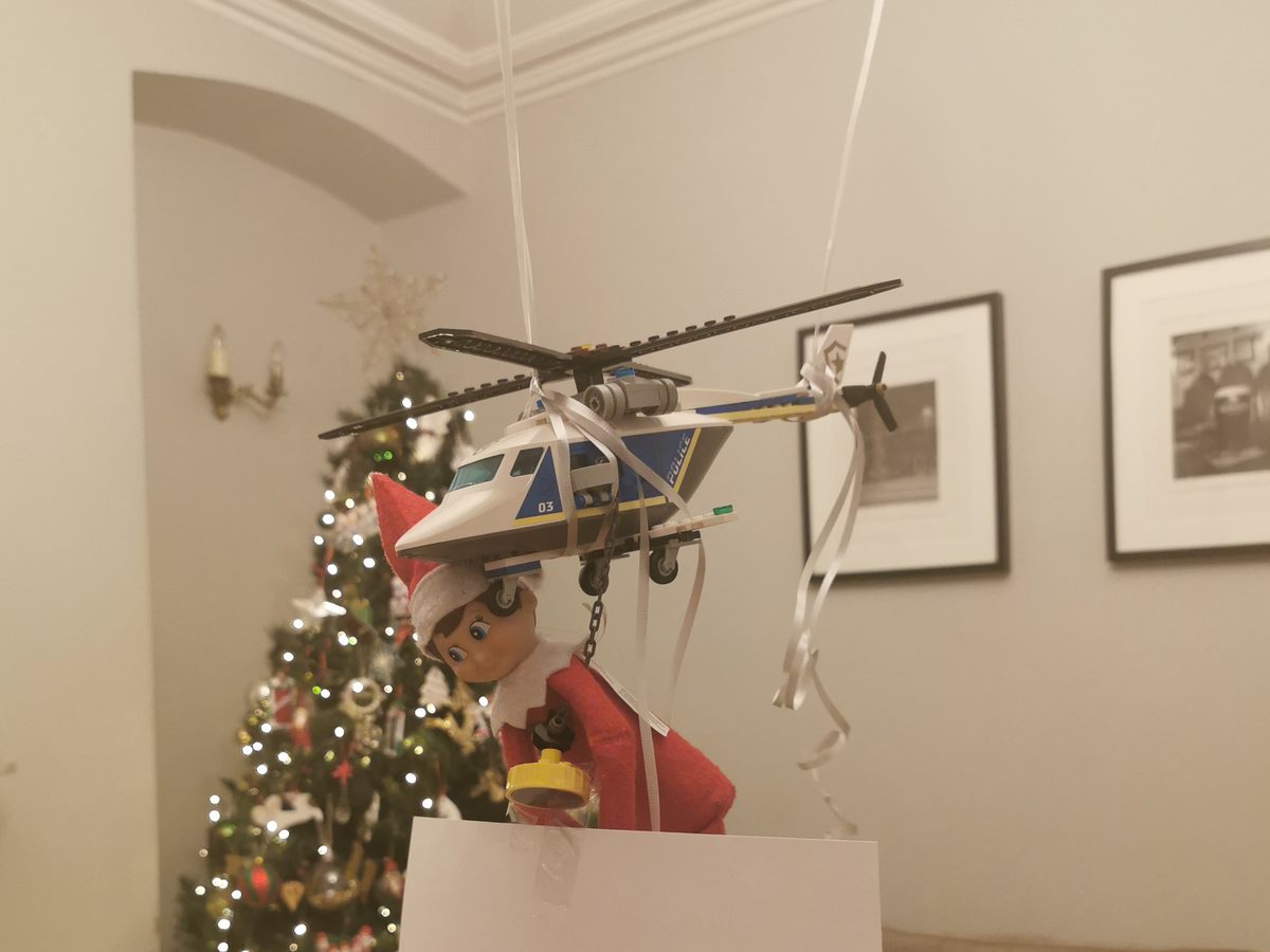 Elf suspended beneath toy helicopter creating dramatic stunt arrival scene.