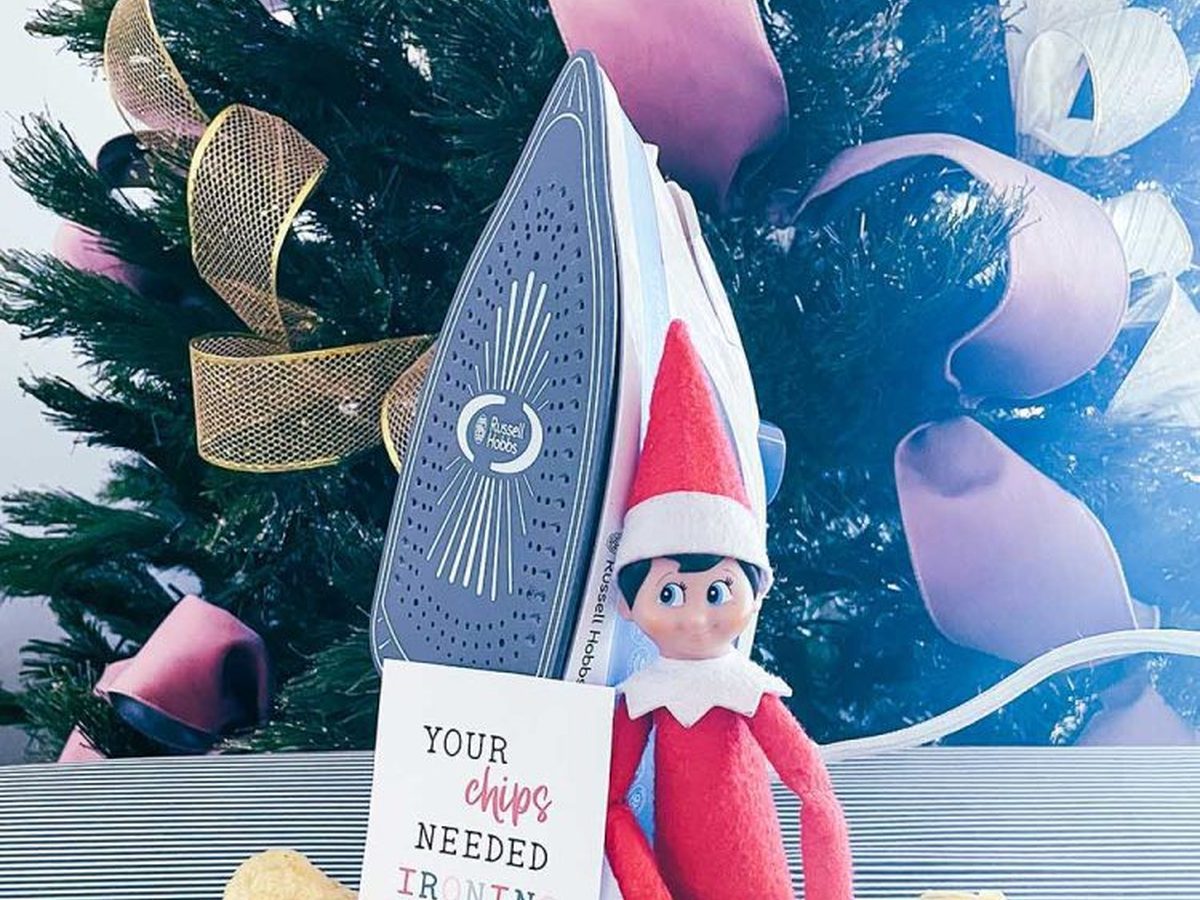 Elf on the shelf ironing chips with toy iron and board on kitchen surface.