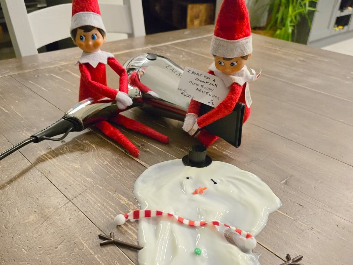 Elf standing beside melted shaving foam snowman with carrot and coal remnants scattered nearby.