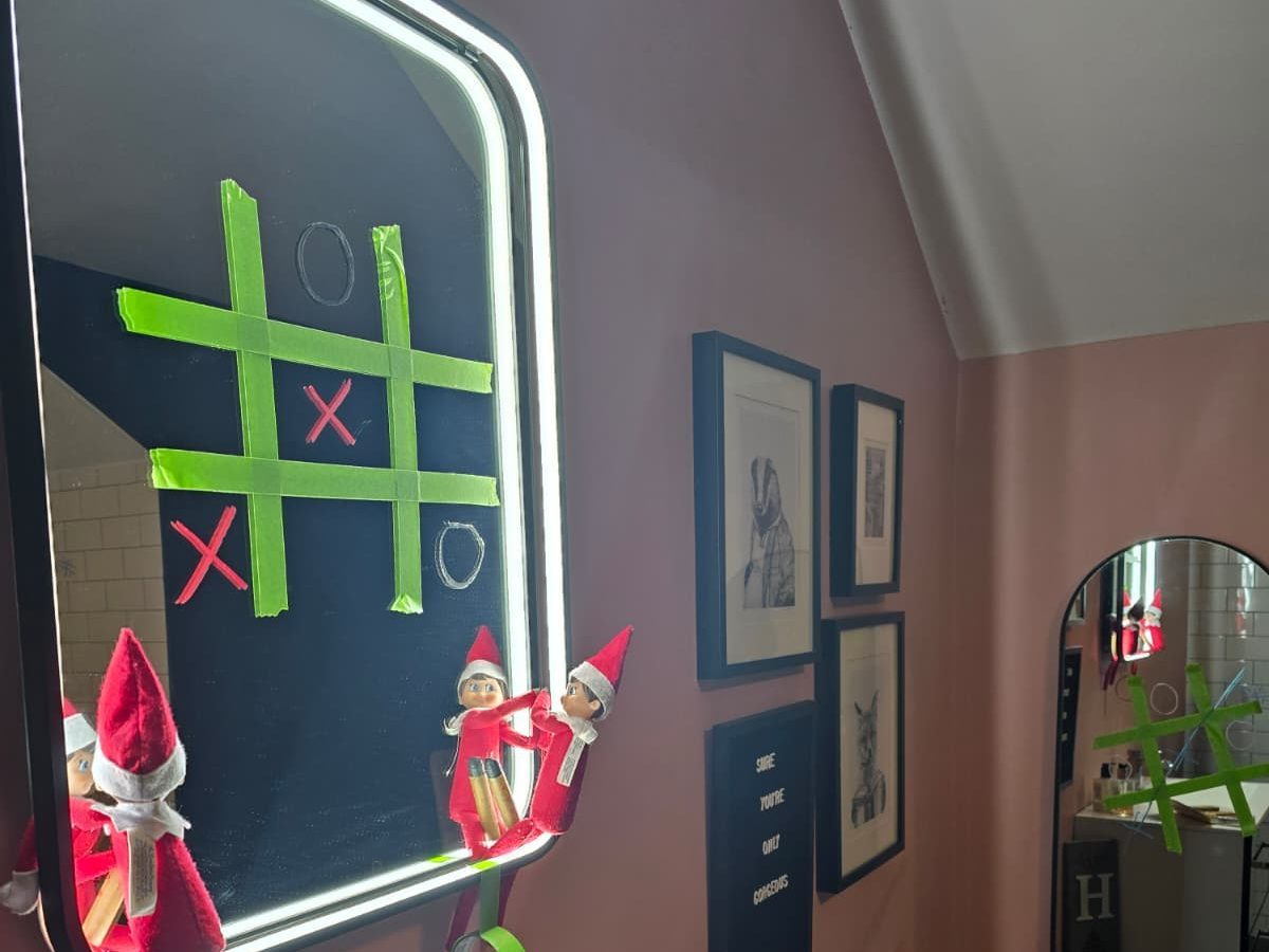 Elf playing tic-tac-toe on bathroom mirror with toy opponent, game in progress.