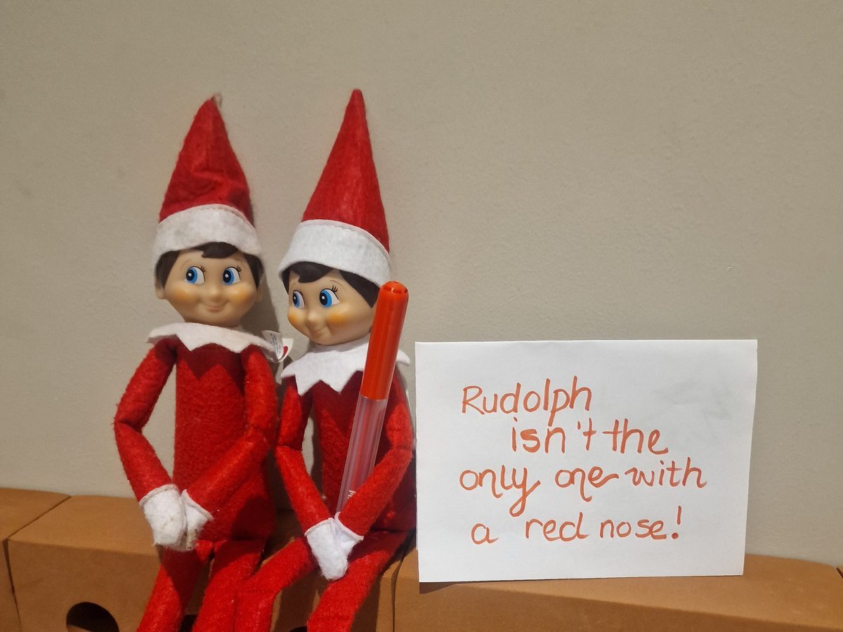 Elf with red marker posed mid-prank on sleeping child's nose.
