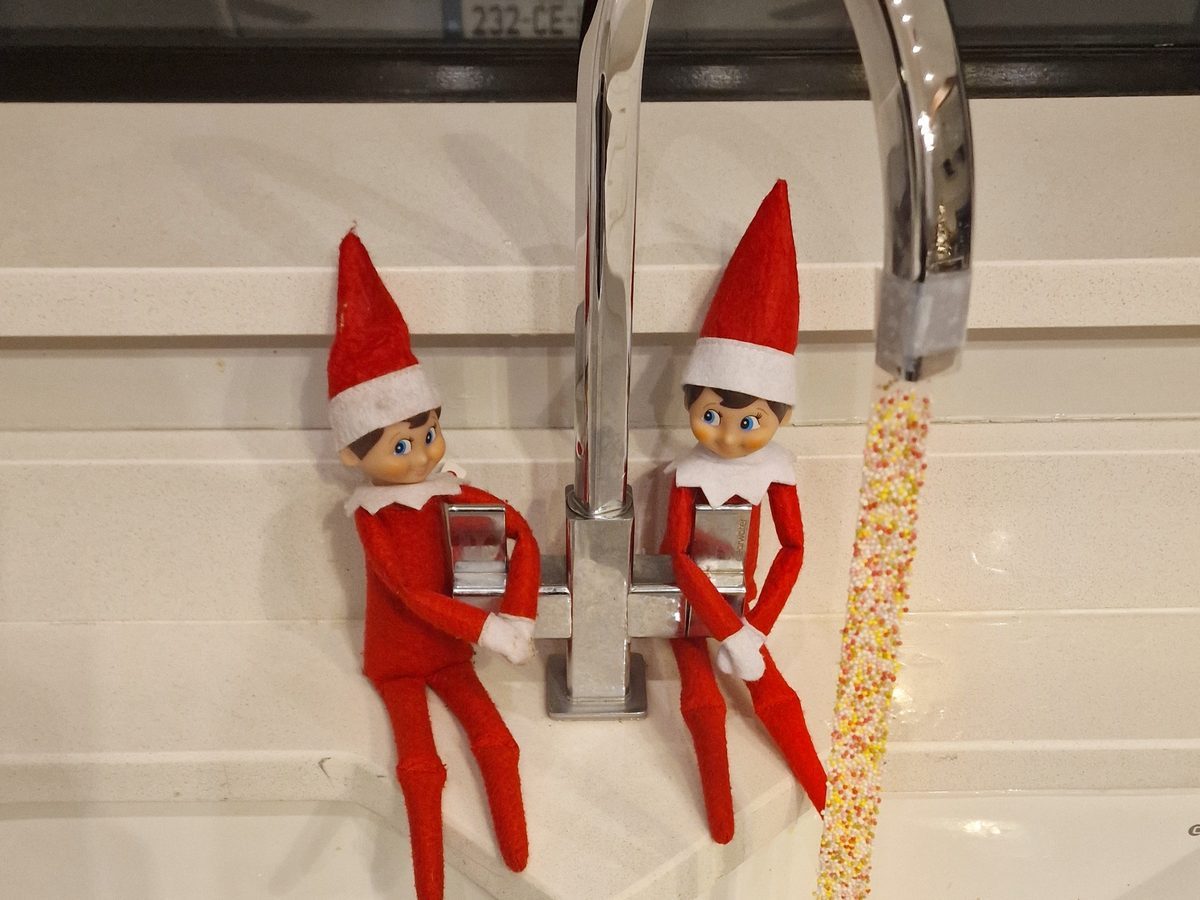 Elf adds colorful sprinkles around kitchen faucet in festive decoration scene.