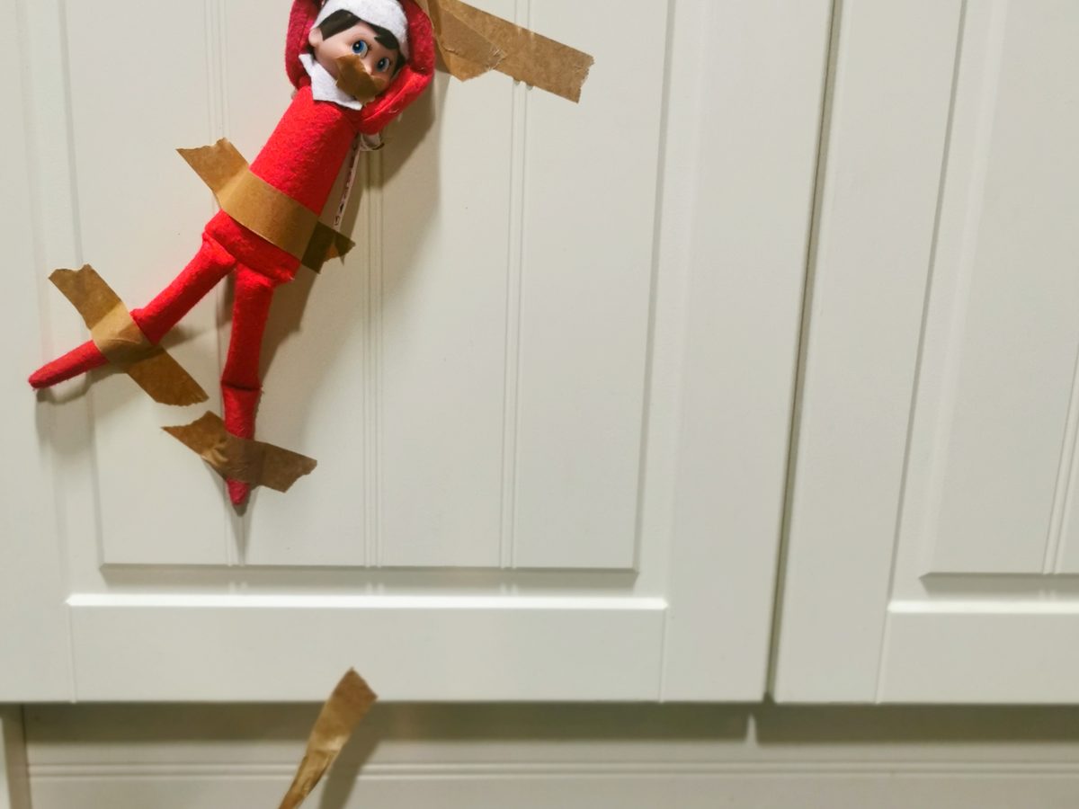 Elf tangled in tape and stuck to wall surrounded by scattered toys.