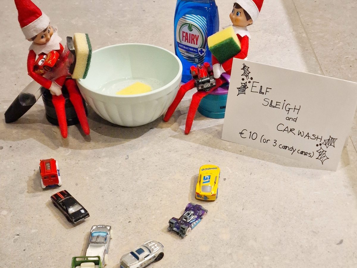 Elf with sponge washing toy cars arranged beside shallow water bowl.